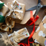 Schere mit Geschenkband und viele in Kraftpapier verpackt Geschenke liegen auf einem Tisch.