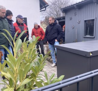 mehrere Menschen stehen im Vorgarten eines Wohnhauses und unterhalten sich über ein Außengerät einer Wärmepumpenanlage, die im Vordergrund steht.