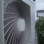 Außengerät einer Luftwärmepumpe, dahinter ein Busch und ein Haus in der Ferne
