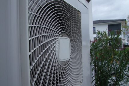 Außengerät einer Luftwärmepumpe, dahinter ein Busch und ein Haus in der Ferne